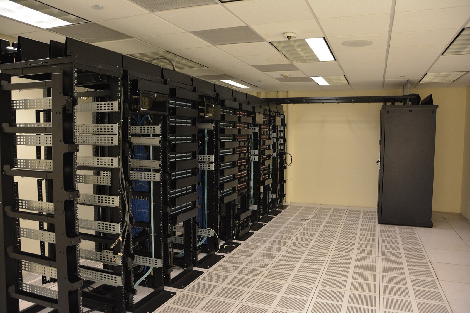 Rows of illuminated server racks inside a data center aisle, the kind of computing infrastructure Applied Digital leases to hyperscalers. Photo: Carl Lender / Wikimedia Commons, CC BY 2.0