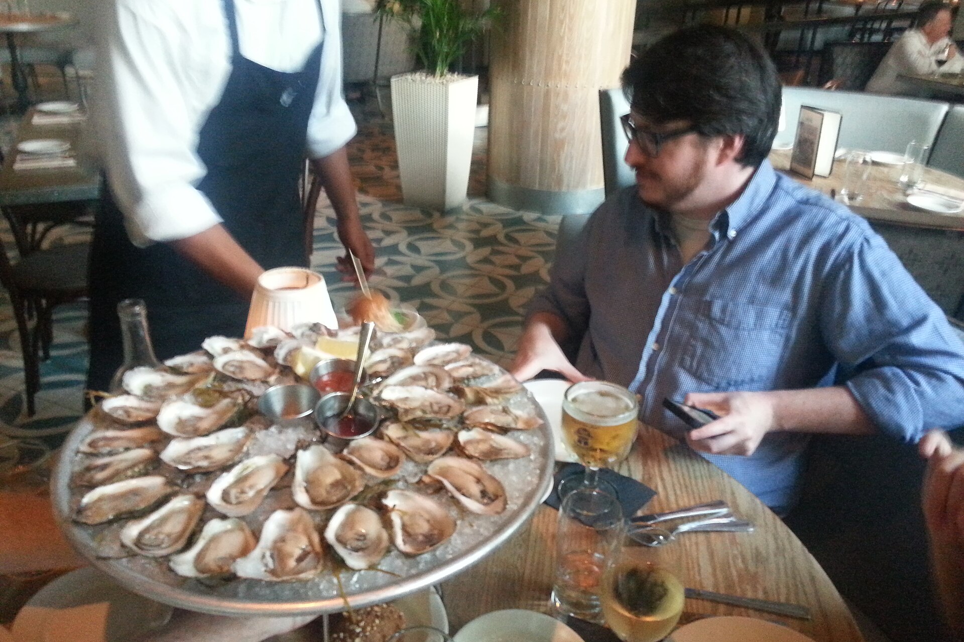 Server delivering a platter of raw oysters on the half shell to a restaurant table, the food route through which many Vibrio vulnificus infections are acquired. Photo: User:Youdontsmellbad / Wikimedia Commons, CC BY-SA 1.0